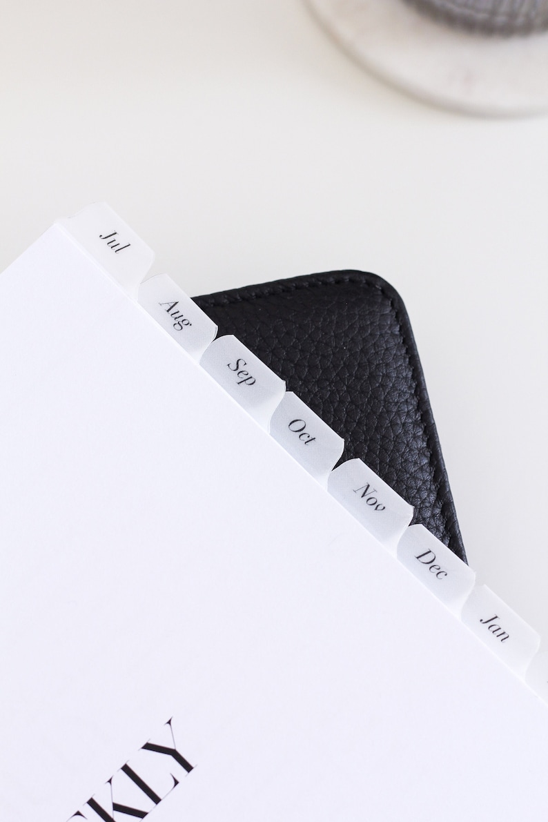 May include: A black leather planner featuring white translucent index tabs labeled with the months Jul, Aug, Sep, Oct, Nov, Dec, and Jan. The planner pages are white with black text, and the cover has a textured, pebbled surface finish.