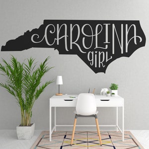 May include: A black outline of the state of North Carolina with the text "Carolina girl" in white lettering. The outline is on a white wall above a white desk with a white chair and a potted plant.