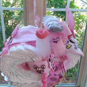 Puede incluir: Un pastel de pa&ntilde;ales rosa y blanco decorado con un tema de Minnie Mouse. El pastel est&aacute; cubierto con un lazo rosa, un calcet&iacute;n blanco y una cabeza de Minnie Mouse rosa y blanca. El pastel est&aacute; rodeado de cintas y lazos rosas y blancos.