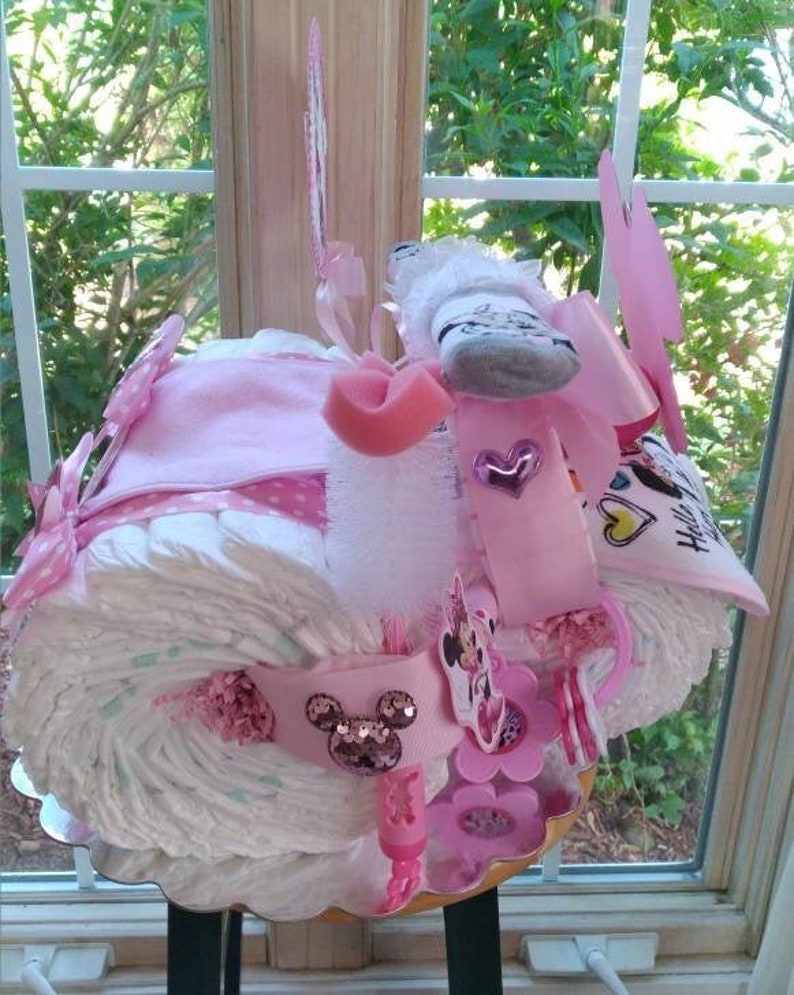Puede incluir: Un pastel de pa&ntilde;ales rosa y blanco decorado con un tema de Minnie Mouse. El pastel est&aacute; cubierto con un lazo rosa, un calcet&iacute;n blanco y una cabeza de Minnie Mouse rosa y blanca. El pastel est&aacute; rodeado de cintas y lazos rosas y blancos.