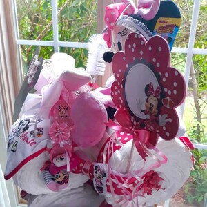 Puede incluir: Un pastel de pa&ntilde;ales rosa y blanco decorado con art&iacute;culos de tem&aacute;tica de Minnie Mouse, incluyendo un juguete de peluche de Minnie Mouse, una flor con lunares y una cara de Minnie Mouse, y una cinta rosa y blanca.