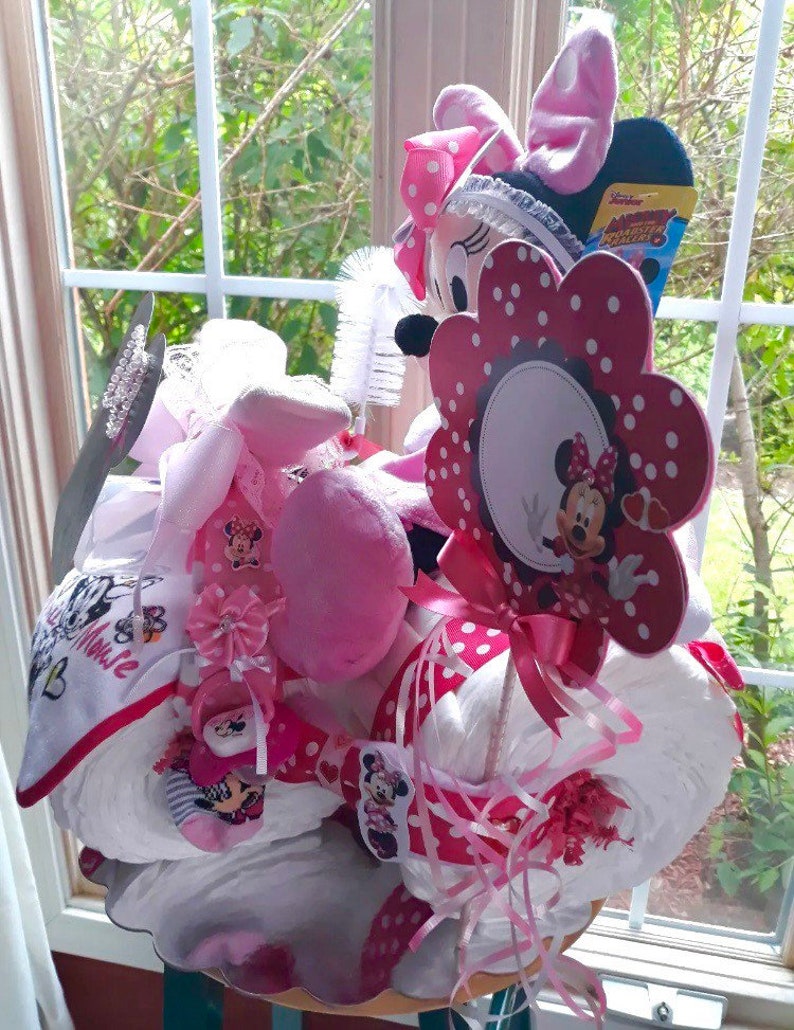 Puede incluir: Un pastel de pa&ntilde;ales rosa y blanco decorado con art&iacute;culos de tem&aacute;tica de Minnie Mouse, incluyendo un juguete de peluche de Minnie Mouse, una flor con lunares y una cara de Minnie Mouse, y una cinta rosa y blanca.