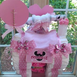 Puede incluir: Pastel de pa&ntilde;ales rosa y blanco decorado con adornos de Minnie Mouse. El pastel est&aacute; cubierto con un lazo rosa y una cinta de lunares rosa y blanco. El pastel tambi&eacute;n est&aacute; decorado con una cinta de lunares rosa y blanco y un lazo de lunares rosa y blanco. El pastel est&aacute; sobre un soporte para pasteles blanco.