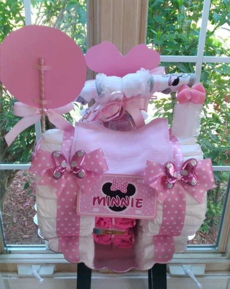 Puede incluir: Pastel de pa&ntilde;ales rosa y blanco decorado con adornos de Minnie Mouse. El pastel est&aacute; cubierto con un lazo rosa y una cinta de lunares rosa y blanco. El pastel tambi&eacute;n est&aacute; decorado con una cinta de lunares rosa y blanco y un lazo de lunares rosa y blanco. El pastel est&aacute; sobre un soporte para pasteles blanco.