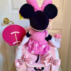 Puede incluir: Un pastel de pa&ntilde;ales Minnie Mouse rosa y negro con un lazo rosa, un vestido de lunares y un cartel rosa y blanco que dice "Minnie".