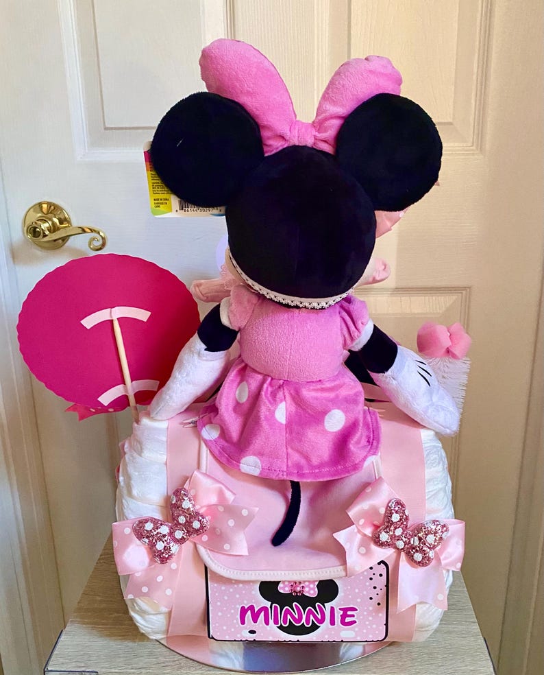 Puede incluir: Un pastel de pa&ntilde;ales Minnie Mouse rosa y negro con un lazo rosa, un vestido de lunares y un cartel rosa y blanco que dice "Minnie".