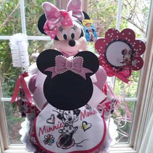 Puede incluir: Un pastel de pa&ntilde;ales Minnie Mouse rosa y negro con un peluche de Minnie Mouse, un babero de lunares rosa y blanco y un cepillo de lunares rosa y blanco.