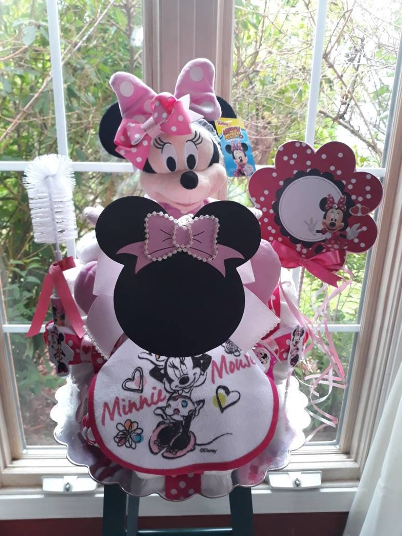Puede incluir: Un pastel de pa&ntilde;ales Minnie Mouse rosa y negro con un peluche de Minnie Mouse, un babero de lunares rosa y blanco y un cepillo de lunares rosa y blanco.