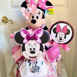 Puede incluir: Un pastel de pa&ntilde;ales Minnie Mouse rosa y negro con un juguete de peluche Minnie Mouse en la parte superior. El pastel est&aacute; decorado con lunares rosas y blancos y lazos. El pastel tambi&eacute;n incluye un cartel tem&aacute;tico de Minnie Mouse que dice "Disney Minnie".