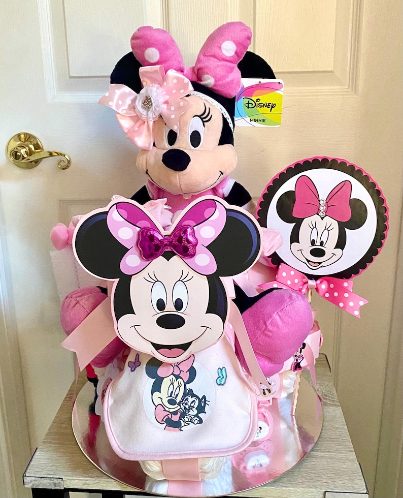 Puede incluir: Un pastel de pa&ntilde;ales Minnie Mouse rosa y negro con un juguete de peluche Minnie Mouse en la parte superior. El pastel est&aacute; decorado con lunares rosas y blancos y lazos. El pastel tambi&eacute;n incluye un cartel tem&aacute;tico de Minnie Mouse que dice "Disney Minnie".