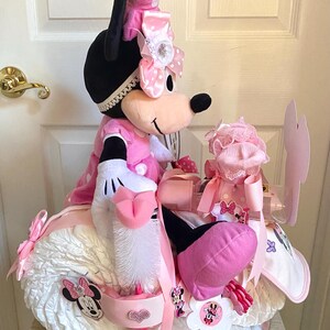 Puede incluir: Un pastel de pa&ntilde;ales rosa y blanco con forma de motocicleta con una mu&ntilde;eca de peluche de Minnie Mouse encima. El pastel est&aacute; decorado con lazos rosas, lunares blancos y aplicaciones de Minnie Mouse.