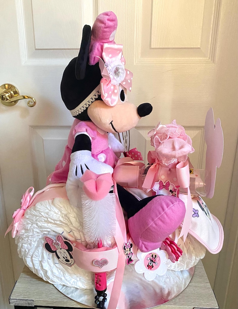 Puede incluir: Un pastel de pa&ntilde;ales rosa y blanco con forma de motocicleta con una mu&ntilde;eca de peluche de Minnie Mouse encima. El pastel est&aacute; decorado con lazos rosas, lunares blancos y aplicaciones de Minnie Mouse.