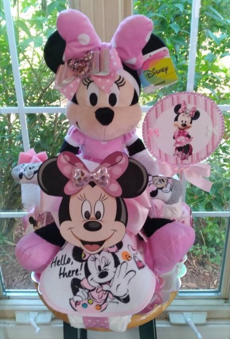 Puede incluir: Un pastel de pa&ntilde;ales Minnie Mouse rosa y blanco con una mu&ntilde;eca de peluche Minnie Mouse en la parte superior. El pastel est&aacute; decorado con lunares rosas y blancos y un lazo rosa. El pastel tambi&eacute;n incluye un letrero que dice "Hello, There!" y una piruleta de Minnie Mouse.