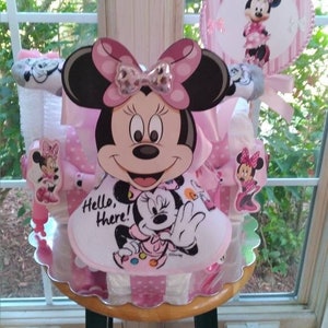 Puede incluir: Un pastel de pa&ntilde;ales rosa y blanco decorado con personajes de Minnie Mouse. El pastel est&aacute; coronado con un gran recorte de cabeza de Minnie Mouse y un babero m&aacute;s peque&ntilde;o de Minnie Mouse. El pastel est&aacute; rodeado de cinta de lunares rosa y blanca.