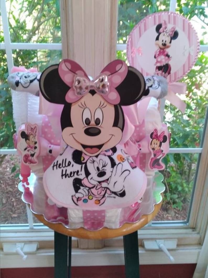 Puede incluir: Un pastel de pa&ntilde;ales rosa y blanco decorado con personajes de Minnie Mouse. El pastel est&aacute; coronado con un gran recorte de cabeza de Minnie Mouse y un babero m&aacute;s peque&ntilde;o de Minnie Mouse. El pastel est&aacute; rodeado de cinta de lunares rosa y blanca.