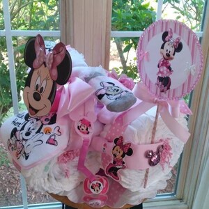 Puede incluir: Un pastel de pa&ntilde;ales rosa y blanco decorado con personajes de Minnie Mouse. El pastel est&aacute; coronado con un c&iacute;rculo rosa y blanco con el tema de Minnie Mouse. El pastel est&aacute; sentado en un taburete de madera.