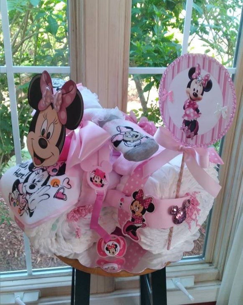 Puede incluir: Un pastel de pa&ntilde;ales rosa y blanco decorado con personajes de Minnie Mouse. El pastel est&aacute; coronado con un c&iacute;rculo rosa y blanco con el tema de Minnie Mouse. El pastel est&aacute; sentado en un taburete de madera.