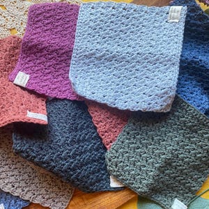 May include: A collection of eight crocheted dishcloths in various colors, including blue, green, pink, and brown. The cloths are made with a textured stitch and are arranged in a pile.