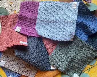 May include: A collection of eight crocheted dishcloths in various colors, including blue, green, pink, and brown. The cloths are made with a textured stitch and are arranged in a pile.