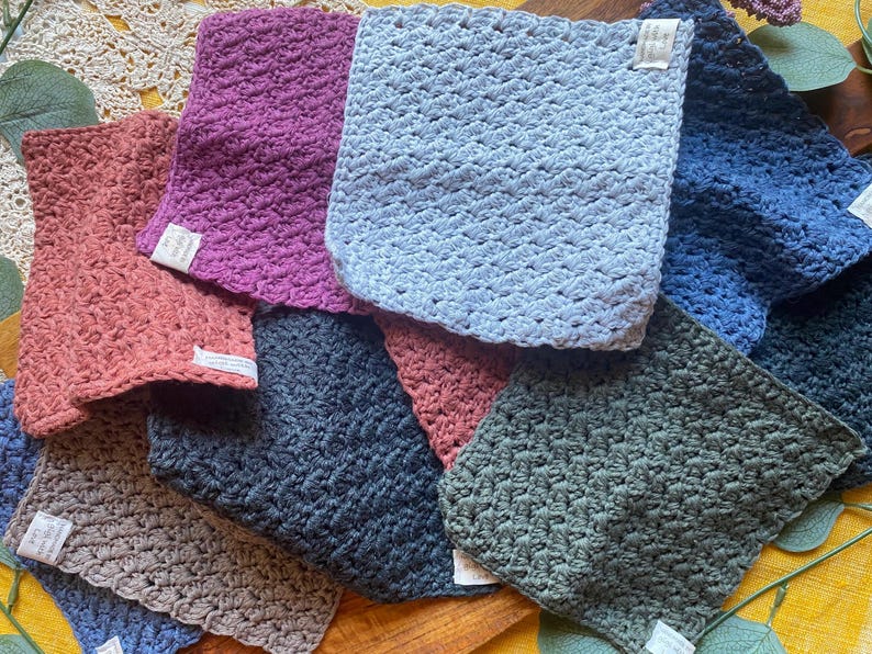 May include: A collection of eight crocheted dishcloths in various colors, including blue, green, pink, and brown. The cloths are made with a textured stitch and are arranged in a pile.