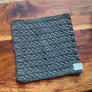 May include: A dark gray crocheted dishcloth or kitchen towel with a textured pattern. The dishcloth has a white label with the text "The Dishcloth Co."