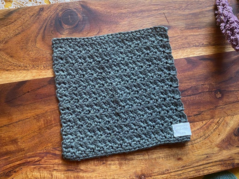 May include: A dark gray crocheted dishcloth or kitchen towel with a textured pattern. The dishcloth has a white label with the text "The Dishcloth Co."