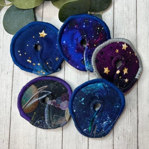 May include: Six fabric circles with a hole in the center. Each circle has a different space-themed print with stars and planets. The circles are blue, purple, and black.