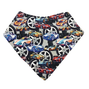 May include: A bib with a repeating pattern of colorful race cars in various colors, including red, blue, yellow, and orange. The cars are set against a background of gray tires and white rims. The bib is shaped like a triangle.