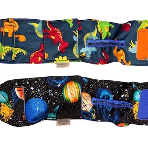 May include: Two fabric wraps with hook and loop fastenings. One has a navy blue background with colourful dinosaurs. The other has a black background with planets, stars, and galaxies. Both have the text "Especially Unique".