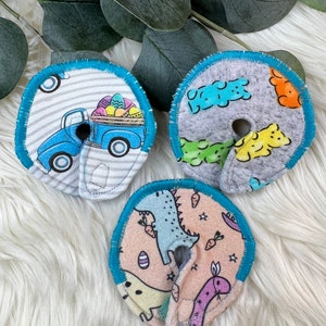 May include: Three round fabric nursing pads with blue trim. The pads feature different designs: a blue truck with Easter eggs, a gray background with colorful gummy bears, and a pink background with dinosaurs and carrots.