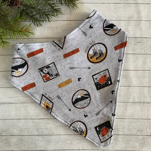 May include: A gray bandana with a repeating pattern of orange and black patches. The patches feature illustrations of camping and hiking scenes with text such as "Hiking Hero", "Epic Explorer", "Fire Starter", "Let's Explore", "Born Leader", "Camp Out", and "Animal Expert".