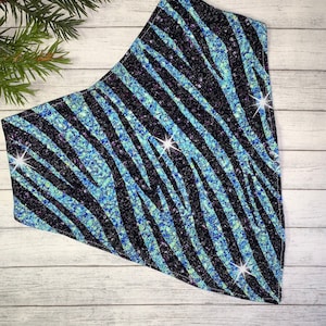 May include: A black and blue patterned bandana with a zebra stripe design. The bandana has a glittery texture and is made of fabric. The background is a light wood surface with a sprig of evergreen.