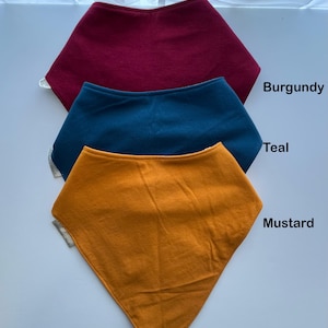 May include: Three colorful baby bibs in burgundy, teal, and mustard yellow. Each bib is a bandana style with a rounded top and a pointed bottom. The colors are labeled with the corresponding color names. The bibs are made of soft fabric.