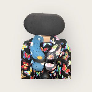 May include: Two colorful neck pillows with butterfly patterns, one with a space theme and the other with a floral theme, are attached to a black wheelchair with a black headrest.