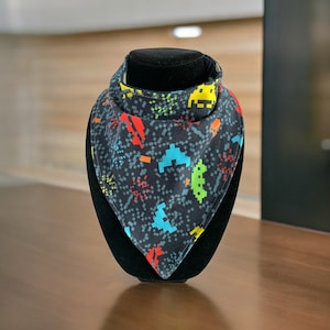 May include: A black bandana with a colourful pixelated design featuring video game characters and fireworks. The bandana is folded and displayed on a black stand.