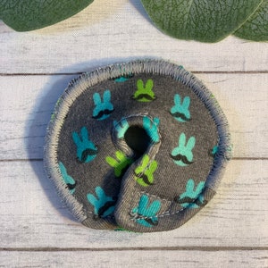 May include: A gray fabric circle with a hole in the center. The fabric has a pattern of blue and green bunnies with black mustaches.