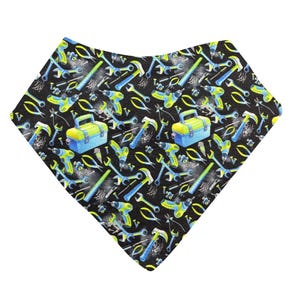 May include: A black bandana-style baby bib featuring a repeating pattern of tools, such as hammers, spanners, and drills. The tools are in blue, yellow, and green, with a blue toolbox. The bib is made of soft fabric.