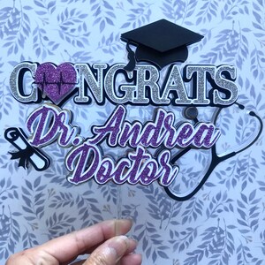 以下が含まれることがあります： 紫と銀のグリッターでできたケーキトッパーで、"Congrats Dr. Andrea Doctor"という文字と、聴診器、卒業帽、心臓に心拍線が描かれたものが付いています。