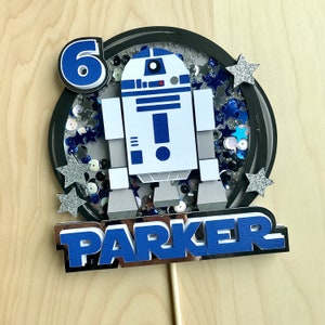 May include: A birthday cake topper featuring a blue and white R2-D2 design with the number 6. The topper includes the name "PARKER" in blue and silver, surrounded by silver glitter stars and blue sequins. It is mounted on a wooden stick.
