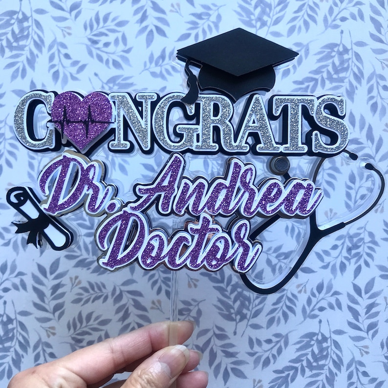 以下が含まれることがあります： 紫と銀のグリッターでできたケーキトッパーで、"Congrats Dr. Andrea Doctor"という文字と、聴診器、卒業帽、心臓に心拍線が描かれたものが付いています。