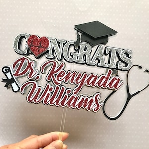 以下が含まれることがあります： 「Congrats Dr. Kenyada Williams」の文字と、卒業帽、聴診器、心臓の鼓動のシンボルが描かれた赤い、白い、銀色のグリッターのケーキトッパーです。