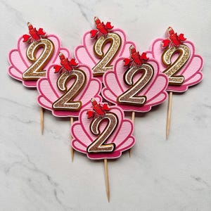 May include: Five cupcake toppers featuring a pink scalloped seashell shape with a glittery gold number two in the center. Each topper is accented with a small red crab character at the top and attached to a thin wooden pick for easy placement.
