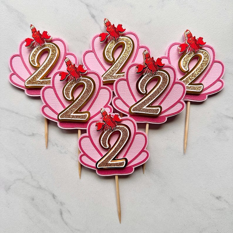 May include: Five cupcake toppers featuring a pink scalloped seashell shape with a glittery gold number two in the center. Each topper is accented with a small red crab character at the top and attached to a thin wooden pick for easy placement.