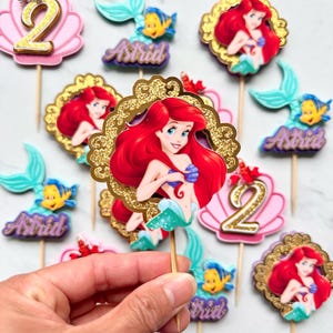 May include: A collection of decorative cupcake toppers featuring The Little Mermaid characters Ariel and Flounder, pink seashells with gold glitter number 2s, and purple name plaques with the name Astrid, all attached to wooden sticks.