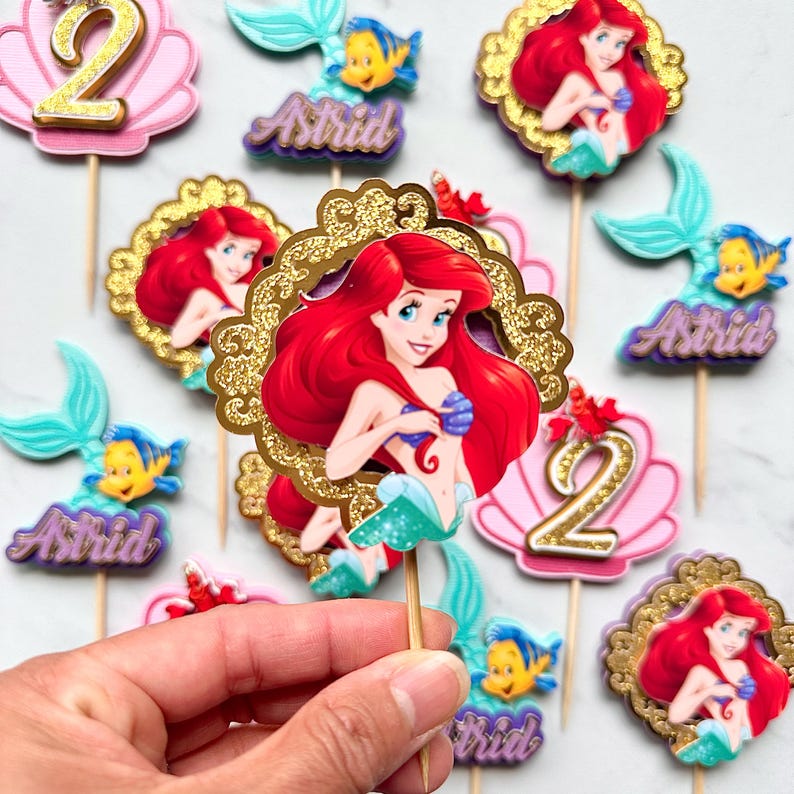 May include: A collection of decorative cupcake toppers featuring The Little Mermaid characters Ariel and Flounder, pink seashells with gold glitter number 2s, and purple name plaques with the name Astrid, all attached to wooden sticks.