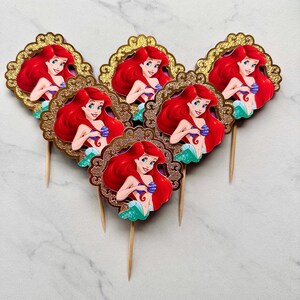 May include: Five cupcake toppers featuring a cartoon mermaid with long red hair and a green tail, each enclosed in a decorative gold glitter frame. The circular cutouts are attached to thin wooden picks, showcasing a character with blue eyes.