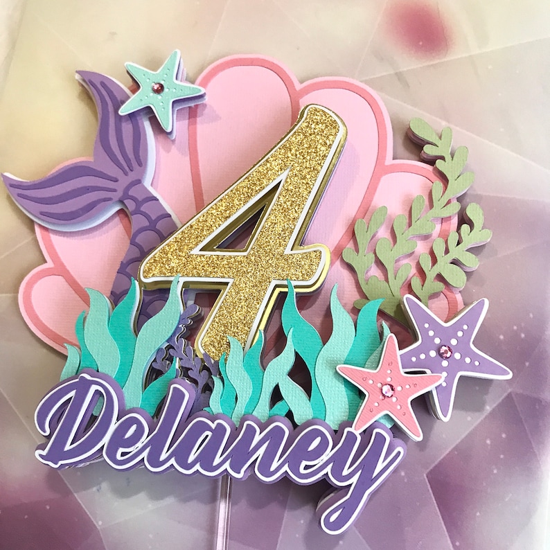 May include: Purple and gold mermaid themed cake topper with the number 4, the name Delaney, and a seashell.
