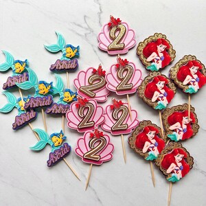 May include: A collection of decorative cupcake toppers featuring various designs: light blue mermaid tails with the name Astrid, pink seashells with a gold glitter number 2 and a small red crab, and circular frames containing a red-haired mermaid.