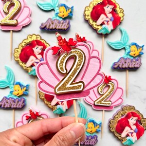 May include: A collection of Little Mermaid themed cake toppers featuring Ariel, Flounder, and Sebastian. The set includes pink scalloped shell cutouts with gold glitter number 2s, circular gold-framed portraits of Ariel, and purple name tags reading Astrid.