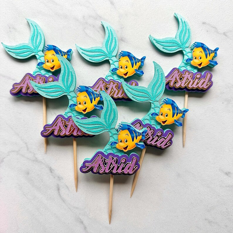 May include: A set of five decorative cake toppers featuring a teal mermaid tail, a yellow and blue fish character, and the name Astrid in purple glittery script. Each topper is attached to a thin wooden pick for insertion into food items.